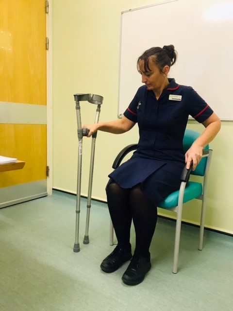 Patient sitting in a chair with one hand on a pair of crutches and their other hand on the arm of the chair.