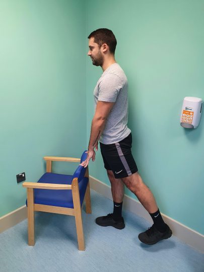 Person who has had a dynamic hip screw operation doing a standing hip extension exercise using a chair for support